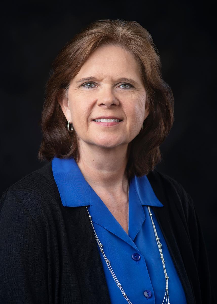 Dr. Susan Seal smiling toward the camera in a blue blouse and black blazer against a black background.